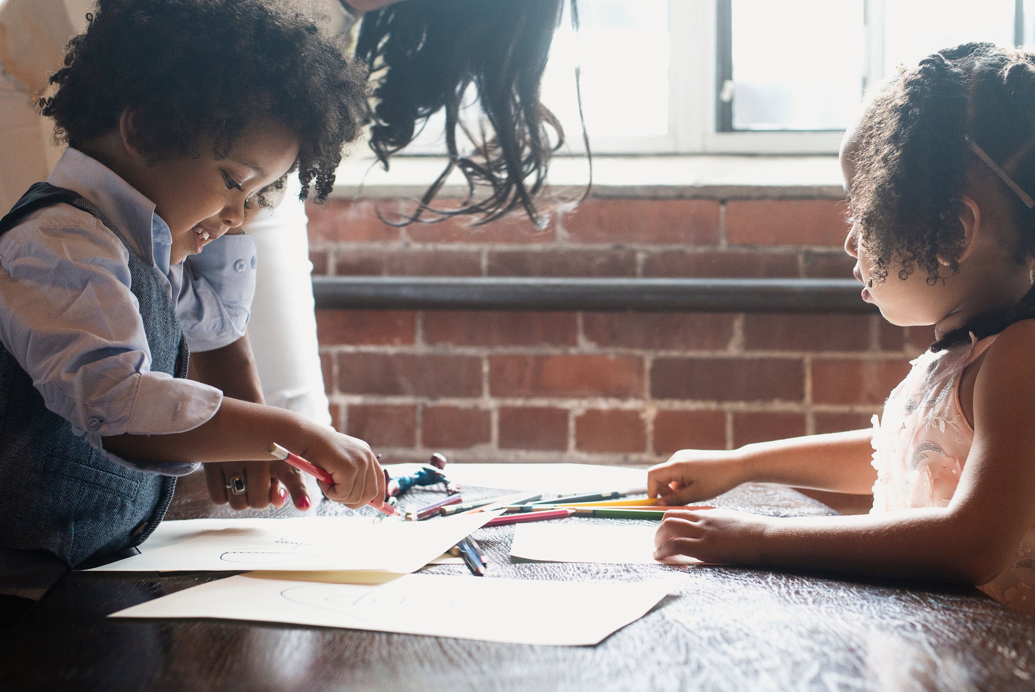 Two children drawing at a table with an adult nearby, in a room with brick walls and large windows.